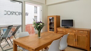 A modern living room with a wooden dining table, white chairs, a TV on a wooden cabinet, and a balcony with a glass door. - Flateli. PDA Vista Mar 3 (Platja d´Aro)