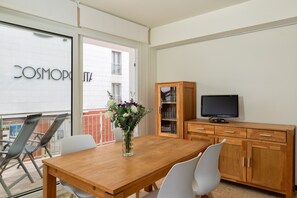 A modern living room with a wooden dining table, white chairs, a TV on a wooden cabinet, and a balcony with a glass door.