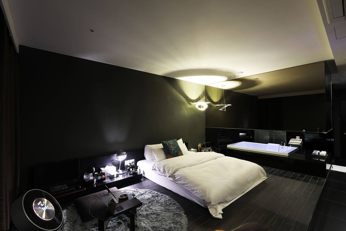 A modern bedroom with a large bed, a vanity with a mirror, a desk with a lamp, and a large window.