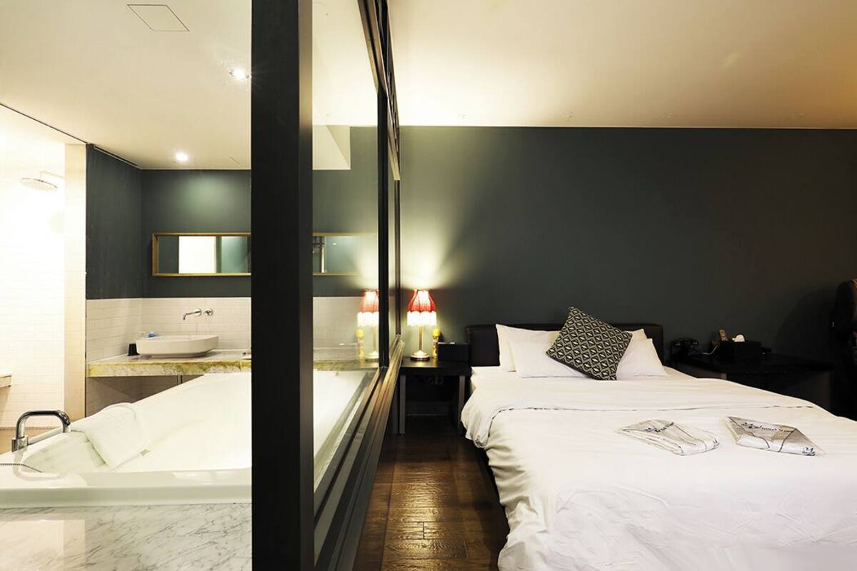 A modern hotel room with a large bed, a separate bathroom with a bathtub, and a nightstand with a lamp.