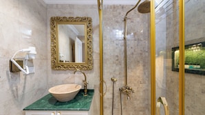 Ein Badezimmer mit Marmorwaschtisch, einem großen Spiegel, einem spiegel mit goldener Rahmung, einer Dusche mit goldener Duschbrause und einer Türklinke aus Gold.