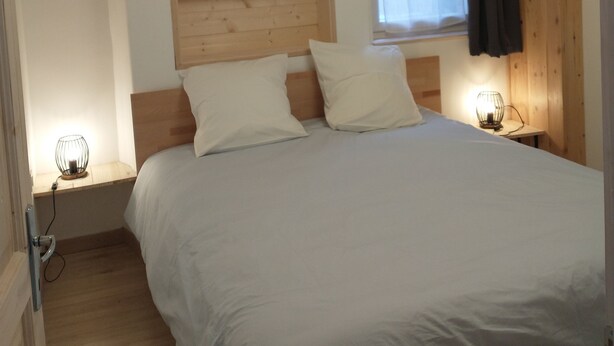 Quarto com cabeceira de madeira, cama com roupa de cama branca, duas luminárias de apoio e uma janela com cortina.