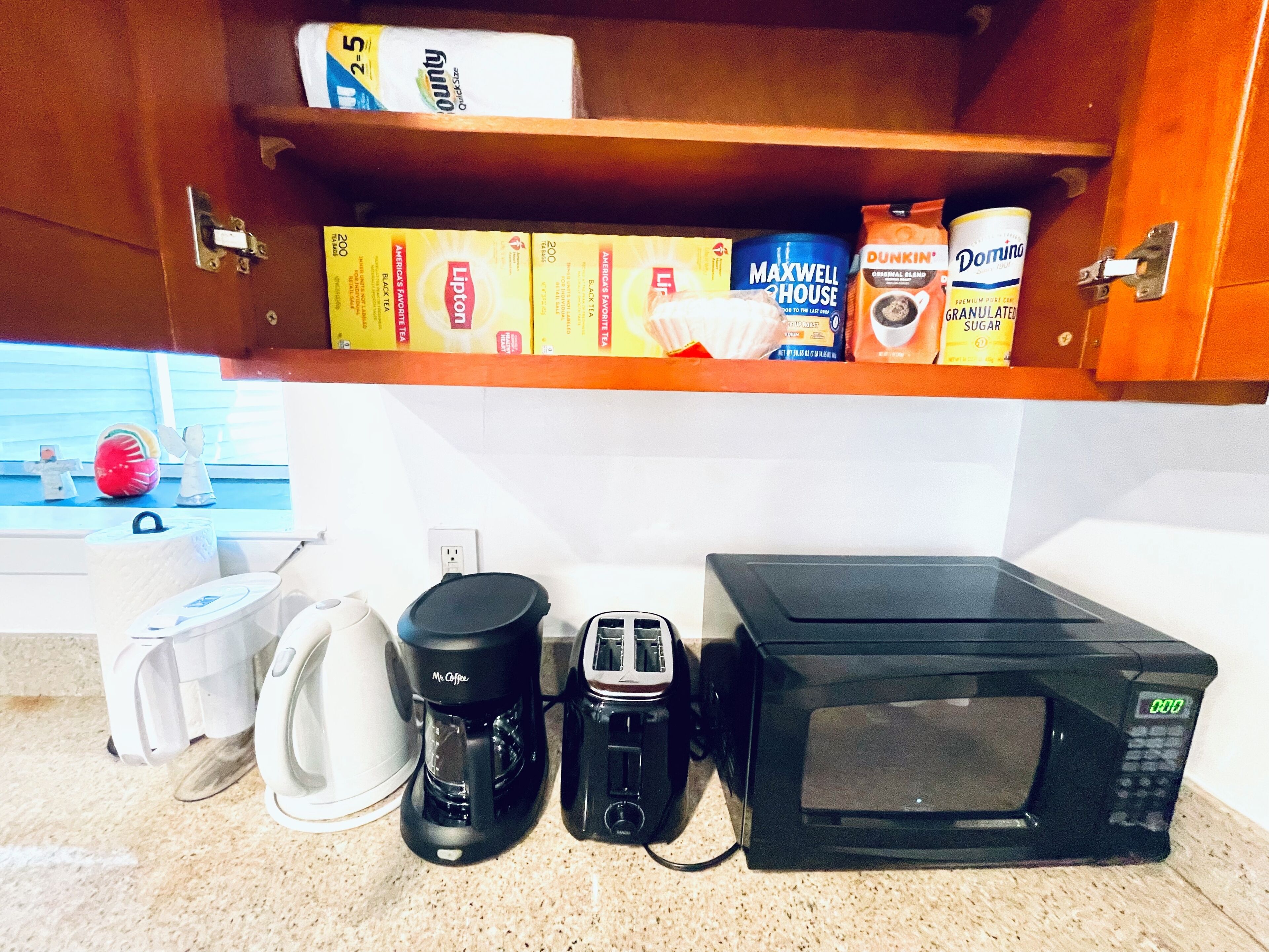 Una cucina con una macchina da caffè, un tostapane e un forno a microonde sul piano di lavoro, e uno scaffale sopra pieno di generi alimentari.