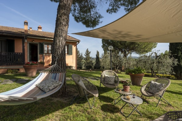 Relax in a serene outdoor space with a hammock, wicker chairs, and a small table, surrounded by lush greenery and mature trees.