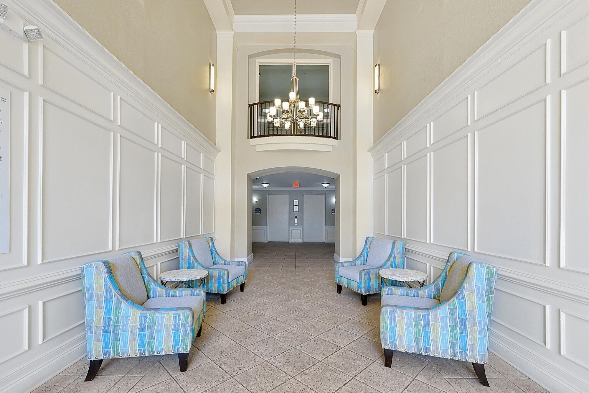 A lobby with a high ceiling, a chandelier, and two sets of blue patterned chairs with small tables.