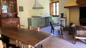 A dining area with a wooden table and chairs, a kitchen with a stove and cabinets, and a window with curtains. - Apartment in Farmhouse (Pelago)