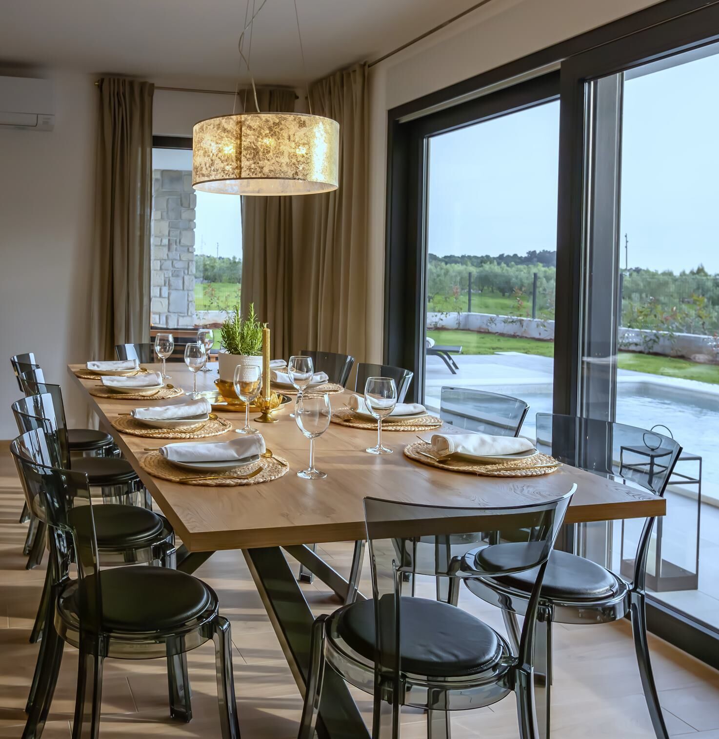 Een eetgedeelte met een lange tafel voor zes personen, omringd door moderne stoelen, een groot raam met uitzicht op groen, en een hanglamp.