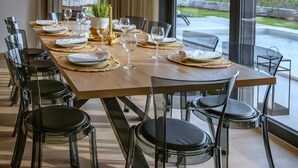Un espace repas avec une longue table dressée pour six personnes, entourée de chaises modernes, une grande fenêtre donnant sur un paysage verdoyant et un luminaire suspendu.