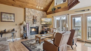A cozy living room with a stone fireplace, leather chairs, a coffee table, and a large window overlooking a deck. - Spacious 4BR Pigeon Forge Cabin with Hot Tub, Game Room, and Mountain Views (Pigeon Forge)