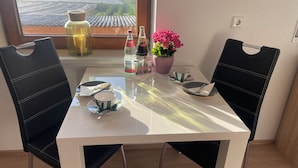 Une table à manger avec des chaises, une vue sur le paysage par la fenêtre, une plante en pot et quelques bouteilles.