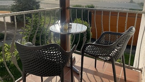 Un balcon avec une table ronde et deux chaises, donnant sur un paysage brumeux où s’étendent des rangées de cultures.