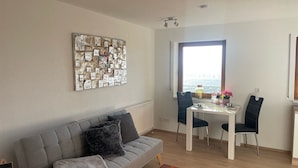 Un salon moderne avec un canapé gris, une table à manger avec des chaises, une grande fenêtre et une œuvre d’art murale.