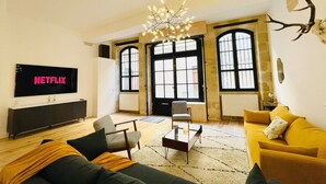 A spacious living room with a large TV displaying 'NETFLIX', a modern chandelier, and a large window with a view. - Loft Bordelais (Bordeaux)