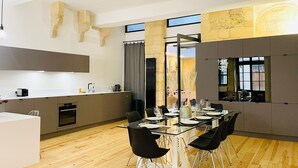 A modern kitchen with a dining area, featuring a glass table and black chairs, wooden flooring, and a large window. - Loft Bordelais (Bordeaux)