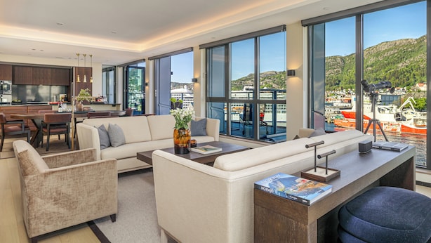 Ein modernes Wohnzimmer mit einer Couch, Sesseln und einem Couchtisch. Ein Küchenbereich mit Schränken und einem Esstisch ist ebenfalls vorhanden. Große Fenster bieten Blick auf Boote und eine Berglandschaft.