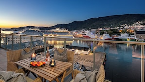 Ein Balkon mit Korbmöbeln, einem Tisch mit Früchten und Kerzen sowie Blick auf Boote und Gebäude über dem Wasser.