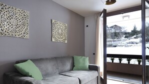 A grey sofa with green cushions, a decorative wall panel, and a view of a snowy landscape through a glass door. - Cornone Di Blumone A5 by Halldis (Ponte Di Legno)