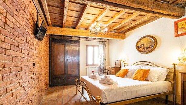 A room with a brick wall, wooden ceiling beams, a bed with white linens and orange pillows, a wooden cabinet, and a wall-mounted mirror.