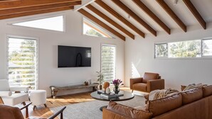 Un vaste salon doté d’un haut plafond, de poutres apparentes, de grandes fenêtres et d’un téléviseur à écran plat fixé au mur.