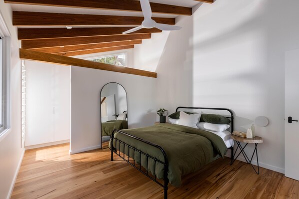 A bedroom with a bed, wooden beams, a ceiling fan, a small table, and a mirror.