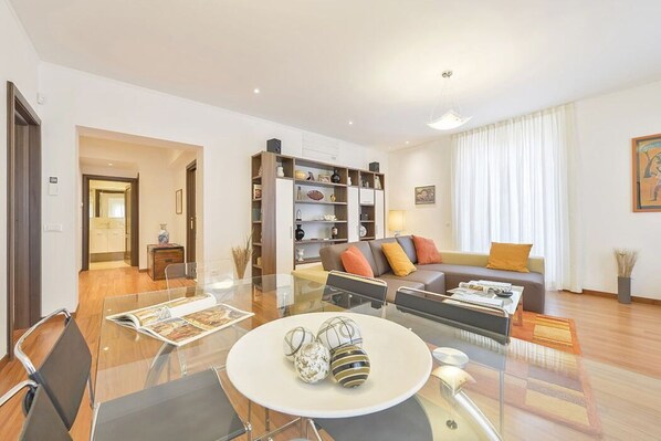 A modern living room with a glass dining table, a sofa, and a bookshelf.