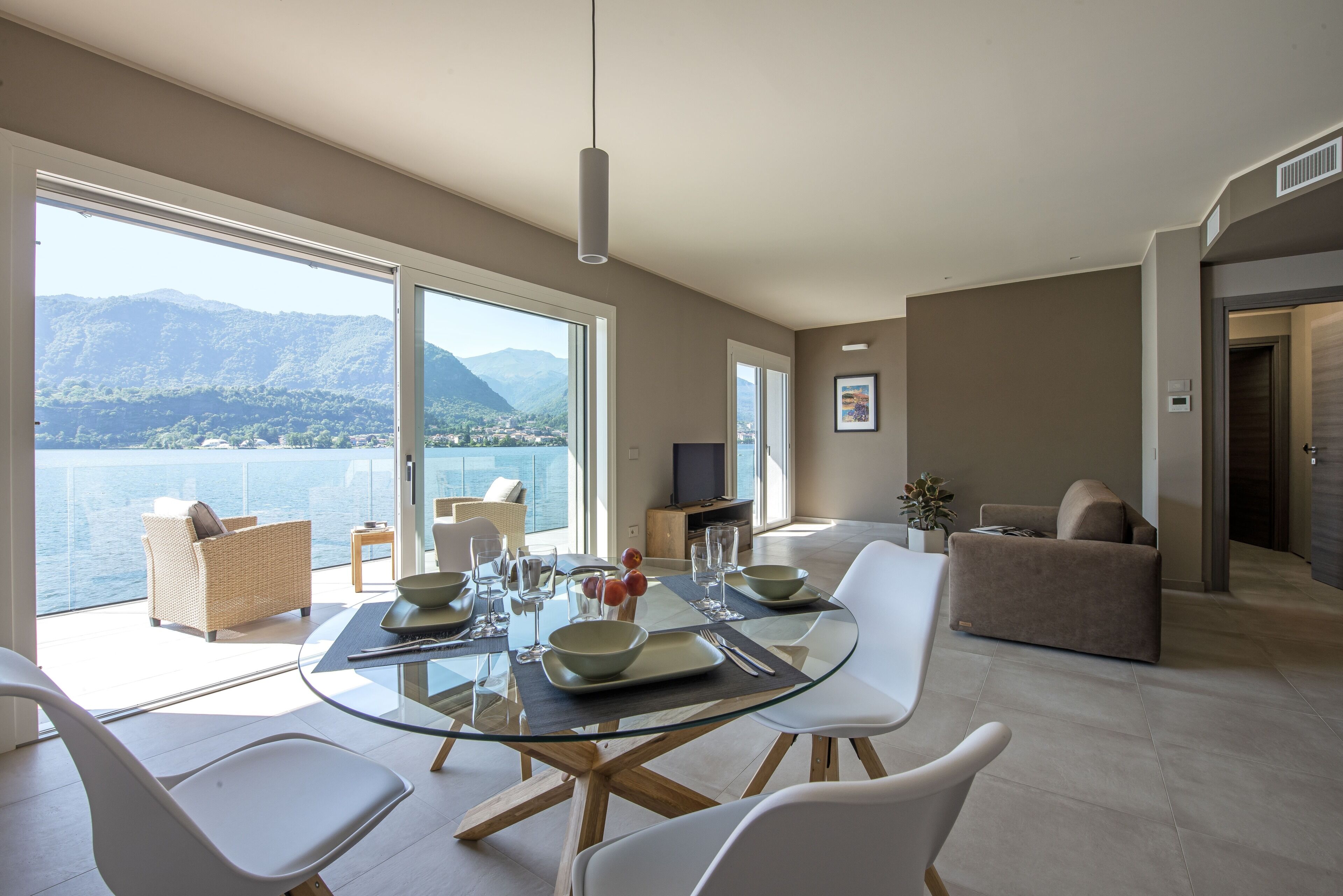 Un espace repas moderne avec une table en verre, des chaises blanches et une vue sur un lac et des montagnes.