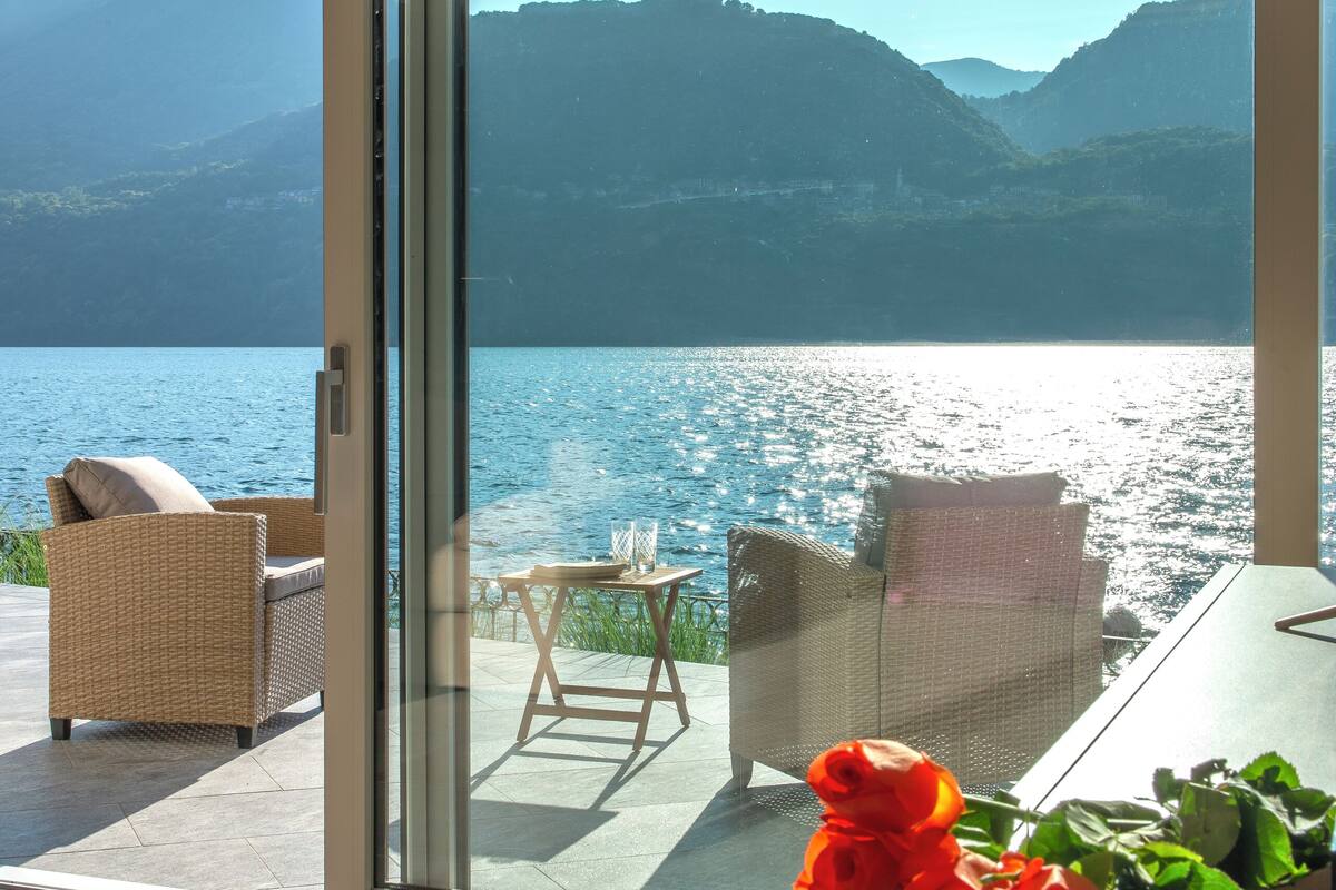 A room with a sliding glass door leading to a balcony overlooking a lake and mountains.