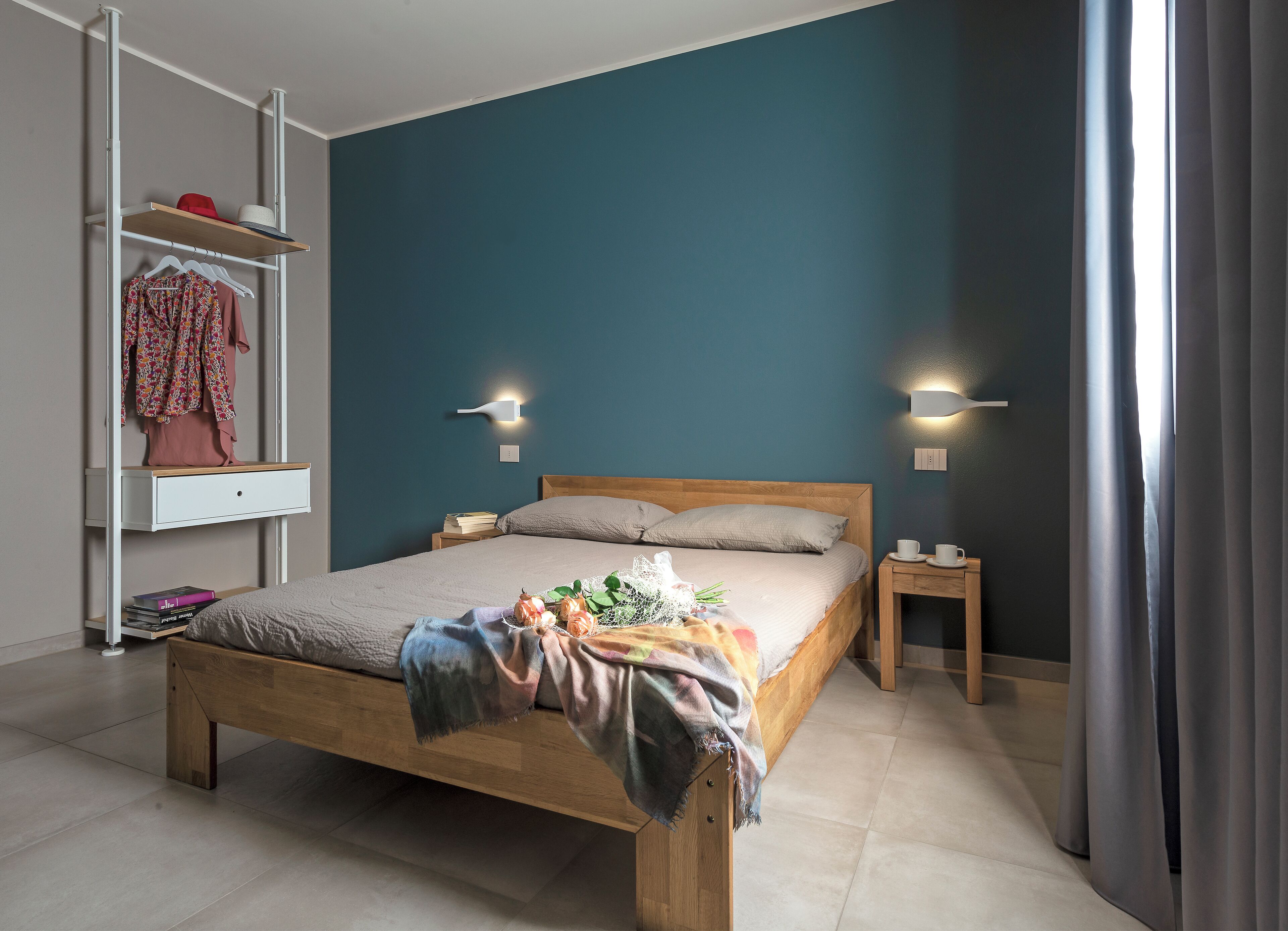 Quarto com cama de madeira, mesa de cabeceira, prateleira com roupas e duas luzes fixadas na parede.