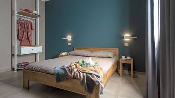Quarto com cama de madeira, mesa de cabeceira, prateleira com roupas e duas luzes fixadas na parede.