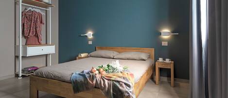 Un dormitorio con una cama de madera, una mesita de noche, un estante con ropa y dos luces fijadas a la pared.