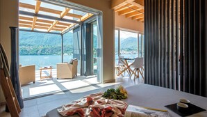 A modern bedroom with a large bed, a view of a lake and mountains, and a balcony with wicker furniture. - d'Ocalée Holiday Experience (Omegna)