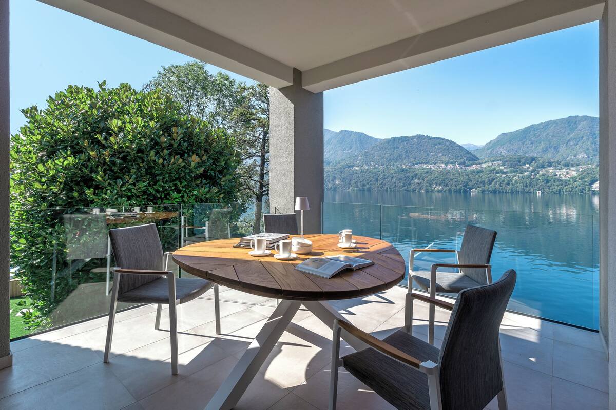 A round table with four chairs, a book, and a cup on a balcony overlooking a lake with mountains in the background.