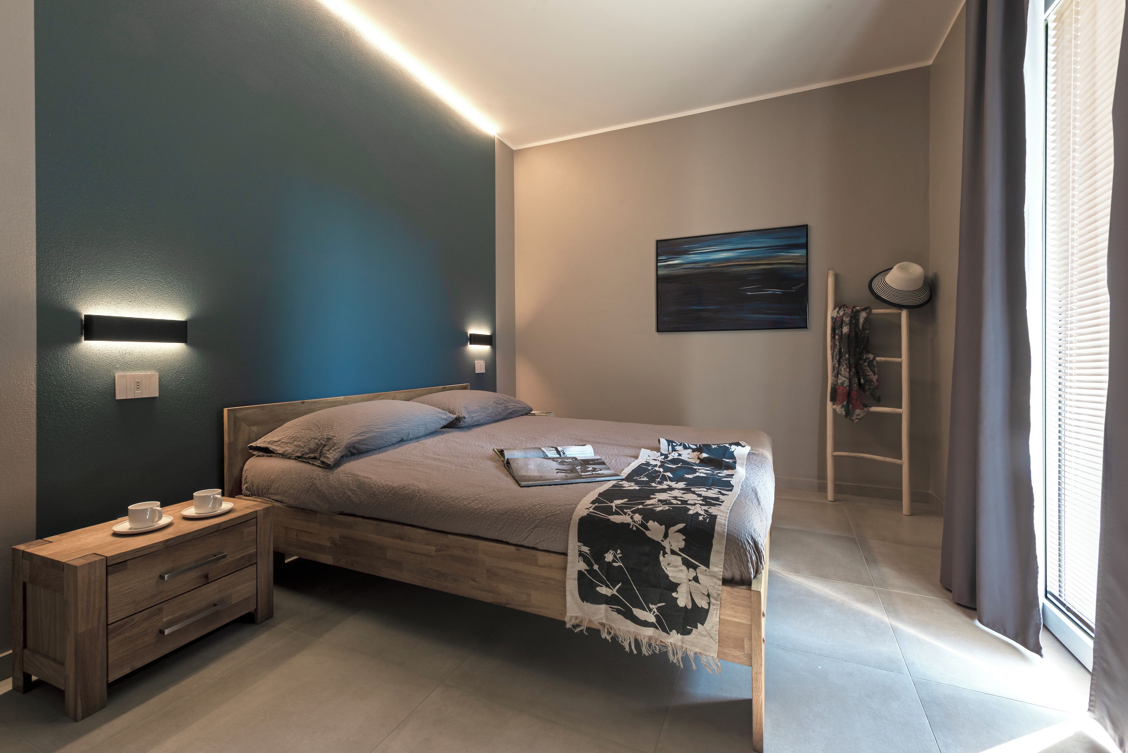 A modern bedroom with a wooden bed, bedside tables, a nightstand, a painting on the wall, and a window with curtains.