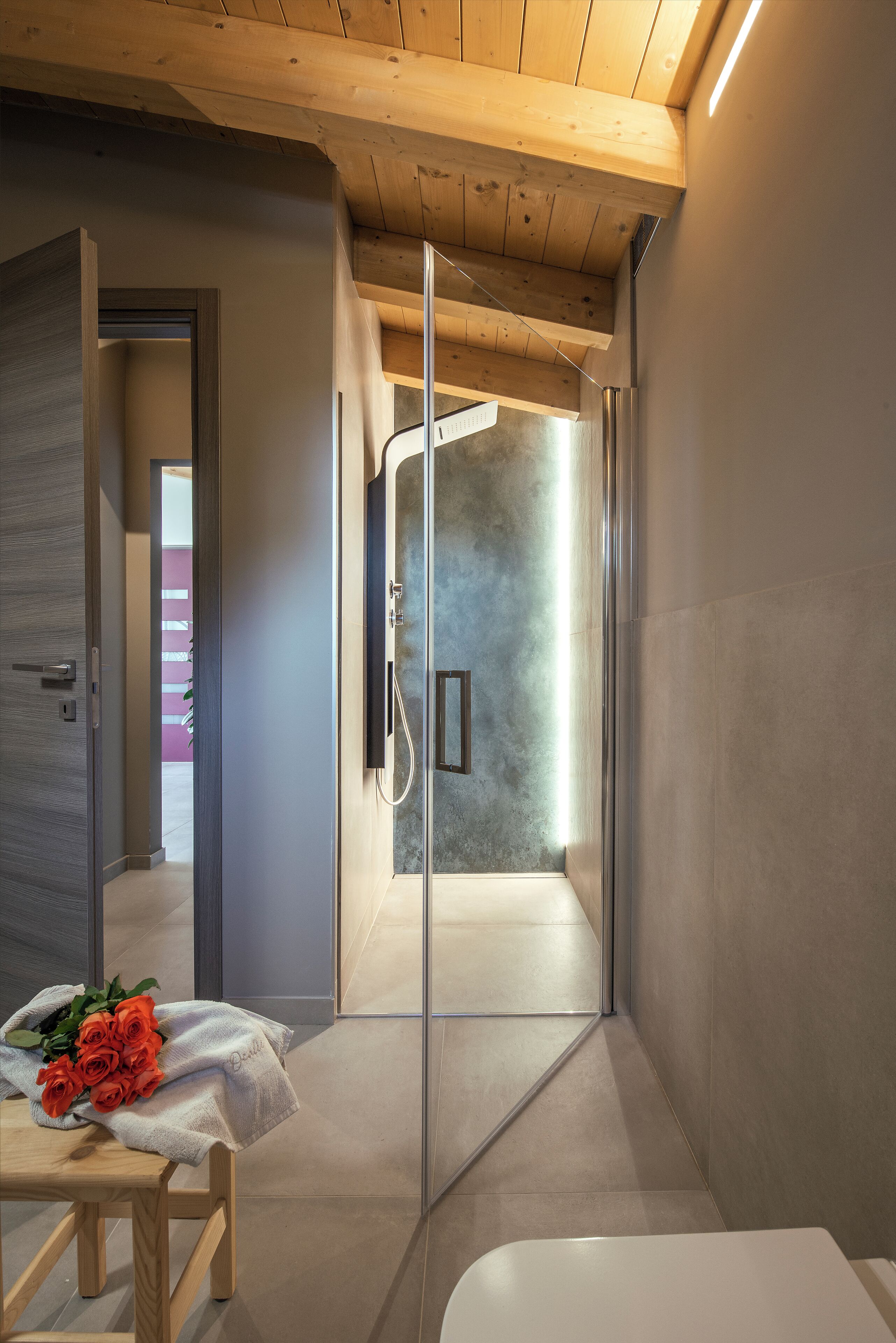 Un baño moderno con ducha, un banco de madera con toalla y rosas, y vista a un edificio rosa en el exterior.