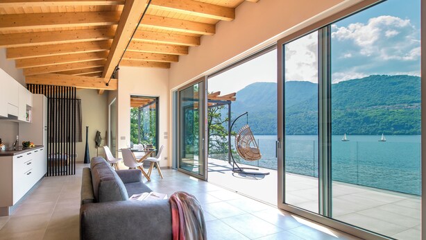 A modern living room with a large sliding glass door leading to a balcony overlooking a lake with sailboats. - d'Ocalée Holiday Experience (Omegna)