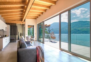 A modern living room with a large sliding glass door leading to a balcony overlooking a lake with sailboats. - d'Ocalée Holiday Experience (Omegna)