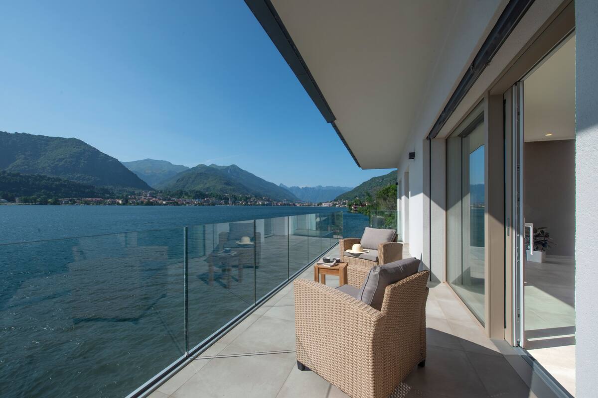 A balcony with a wicker sofa, a small table, and a glass railing overlooking a lake and mountains.