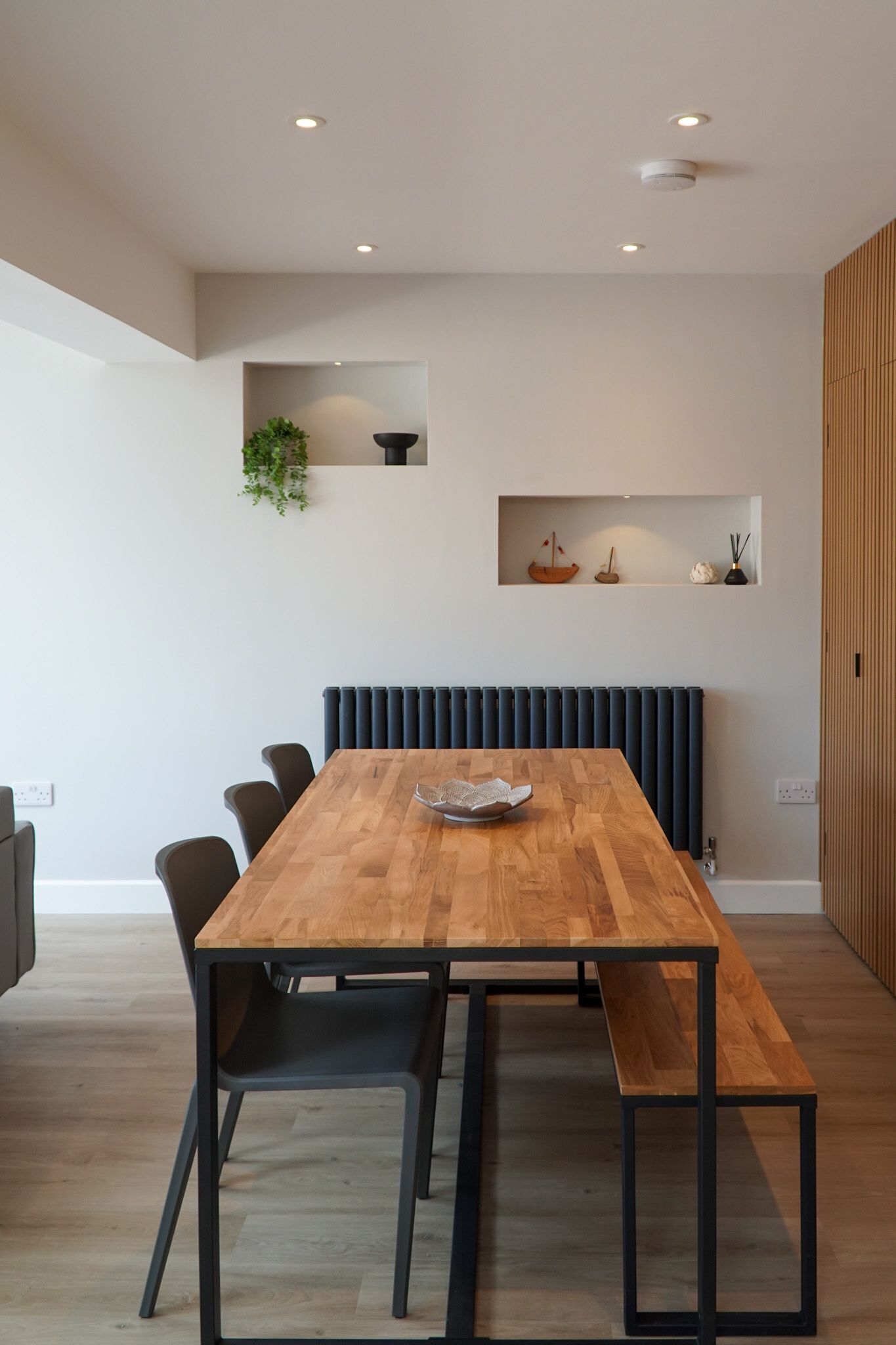 Un espace repas moderne avec une table en bois, des chaises noires et un banc intégré. On y trouve une étagère fixée au mur, ornée d’objets décoratifs et d’une plante.