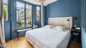 A bedroom with a large bed, a bench, and a view of buildings through large windows. - Industry Chic, by Veeve (Paris)