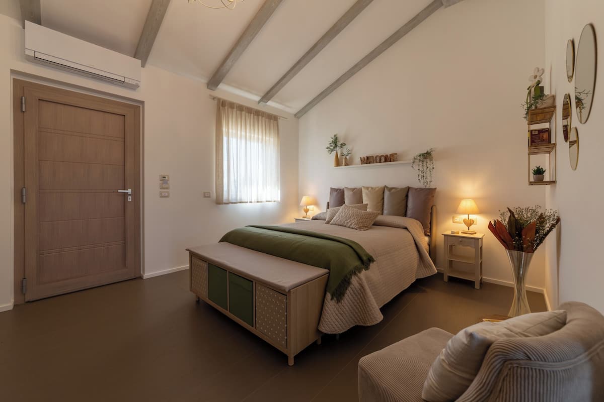 Quarto com cama, mesas de cabeceira, um banquinho e prateleira na parede com plantas e um espelho.