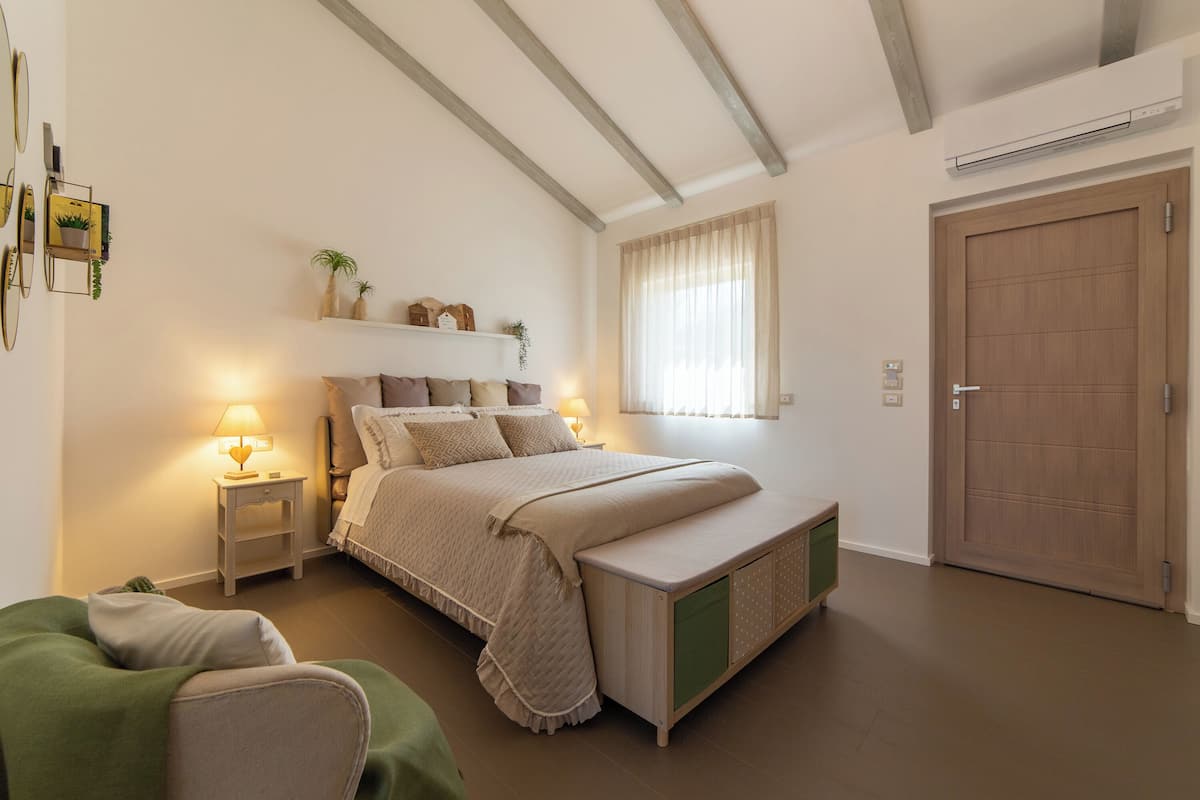 Um quarto com cama, mesas de cabeceira, um criado-mudo, janela com cortinas, porta e prateleira na parede com objetos decorativos.