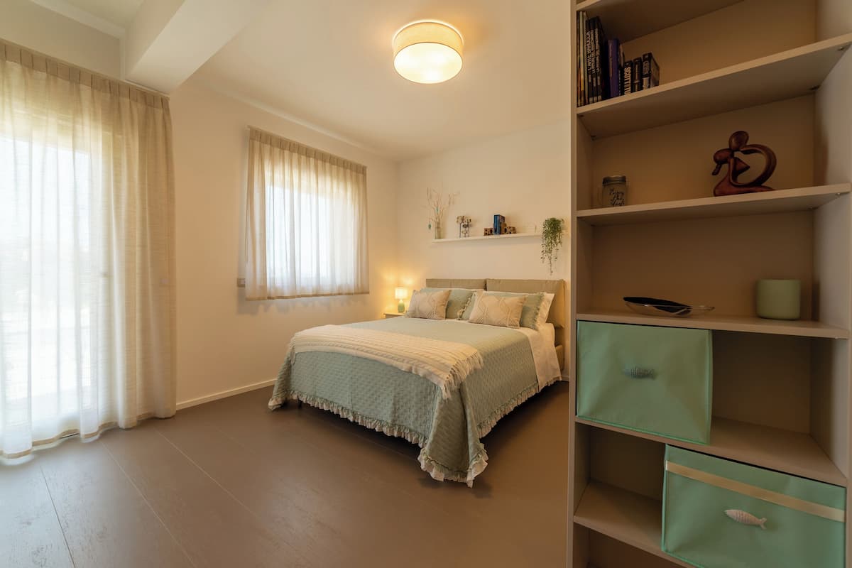 Quarto com cama, mesa de cabeceira, estante com livros e objetos decorativos, e janela com cortinas.