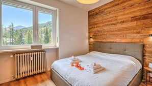 A bedroom with a large window, a wooden wall panel, a bed with towels, and a radiator. - Starkeeper Apartment Ski Base With Balcony Views (Pragelato)