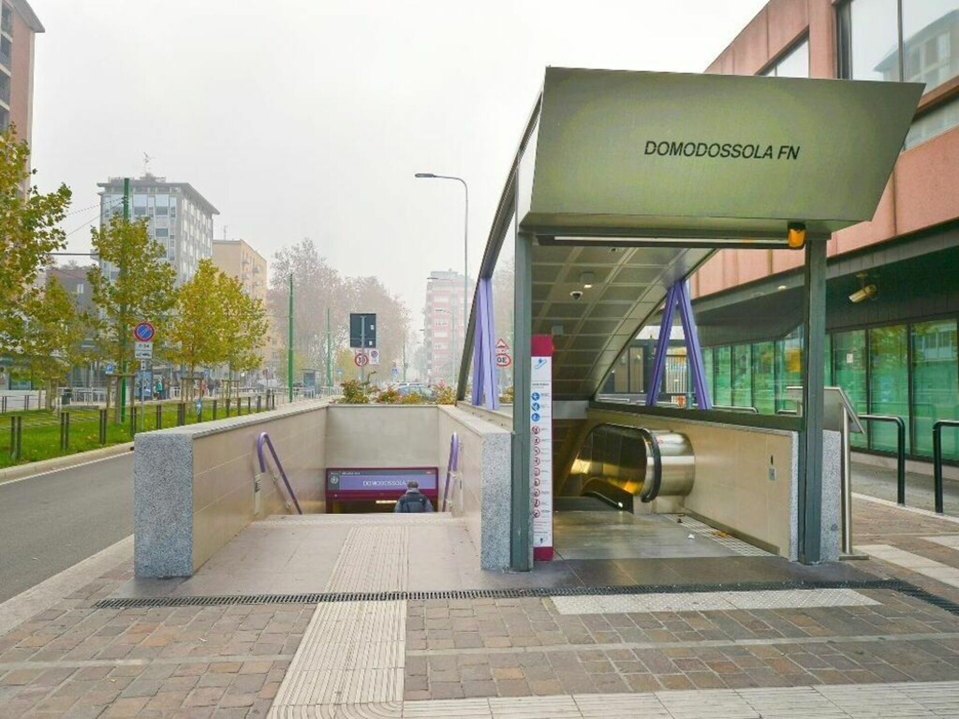 A modern building entrance with a covered waiting area, a ticket machine, and an escalator.