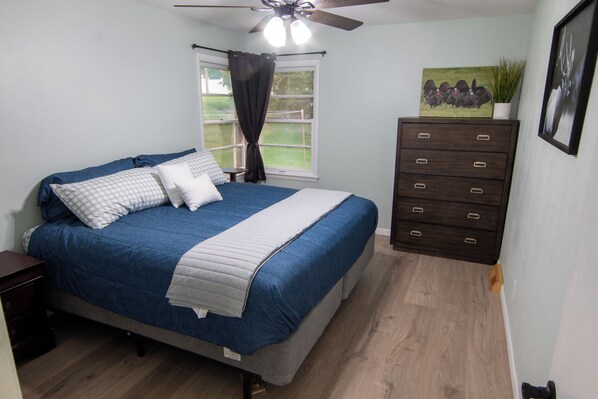 Une chambre à coucher avec un lit, une commode, un ventilateur de plafond, une fenêtre avec des rideaux et un tableau au mur.