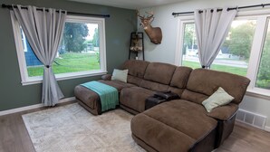 Un salon comprenant un canapé d’angle, une tête de cerf fixée au mur, et une vue sur l’extérieur grâce à deux fenêtres.