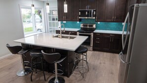 Une cuisine moderne avec des meubles en bois foncé, un réfrigérateur en acier inoxydable, un grand îlot central doté de tabourets de bar et une vue sur l’extérieur grâce à une porte vitrée.