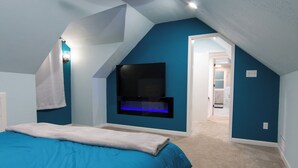 Une chambre avec un plafond en pente, un lit recouvert d’une couverture bleue, un téléviseur fixé au mur et, au fond, une salle de bain visible.