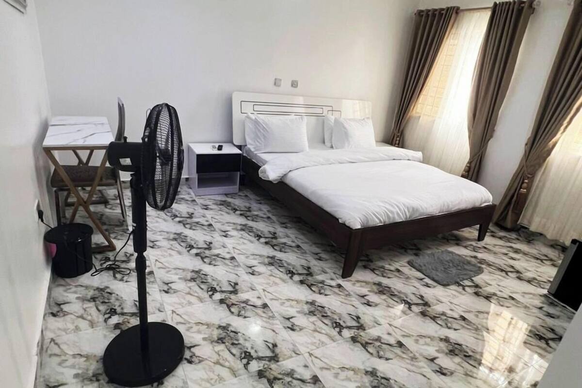 A hotel room with a bed, a desk, a chair, a standing fan, and a marble floor.