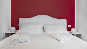 A neatly made bed with white linens and rolled towels, a white headboard, and two bedside tables with white lamps.